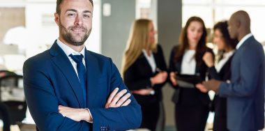 Caucasian businessman leader looking at camera in modern office with multi-ethnic businesspeople working at the background. Teamwork concept. Young man with beard wearing blue suit.