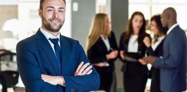 Businessman leader in modern office with businesspeople working Caucasian businessman leader looking at camera in modern office with multi-ethnic businesspeople working at the background. Teamwork concept. Young man with beard wearing blue suit.